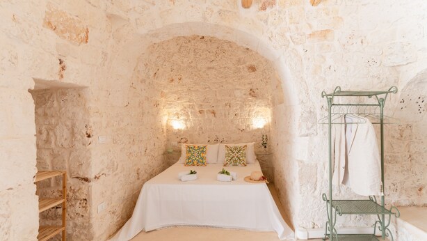 A stone-walled room with a bed, a wooden shelf, and a metal rack.