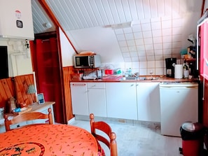 A compact kitchen with a microwave, sink, and refrigerator. There is a dining table with chairs and a patterned tablecloth.