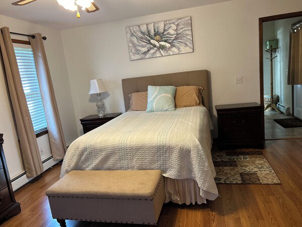 A bedroom with a bed, a nightstand, a lamp, a bench, and a painting on the wall. - Remodeled home near the beach (Dunkirk)