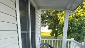 A covered porch with two chairs and a table, white railings, and a view of a green landscape. - Remodeled home near the beach (Dunkirk)