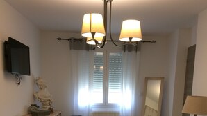 A bedroom with a bed, a chair, a dresser, a television, a window with blinds, and a chandelier. - Guesthouse 'Au Plus Proche Des Vignes' with Private Terrace and Wi-Fi (Montagne)