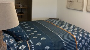 Un dormitorio con cama, lámpara, control remoto, espejo y un cuadro en la pared.