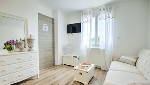Ein Zimmer mit einem weißen Sofa, einer Kommode mit Schubladen, einem an der Wand montierten Fernseher und einem Fenster mit Vorhängen.