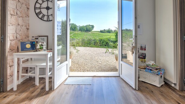 Eine offene Tür, die zu einem Kiesweg mit Blick auf Grünfläche führt. Eine weiße Tafel mit einem gerahmten Zertifikat und eine Wanduhr sind ebenfalls zu sehen.