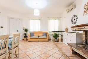Een woonkamer met een bank, een eettafel en stoelen, een keuken met kasten en een haard van baksteen, en een plafondlamp.