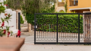 A gated entrance with a metal fence, a stone pillar, and a building with a yellow facade and arched windows. - Villa 'Ivonne's' with Private Terrace, Wi-Fi and Air Conditioning (Villasimius)