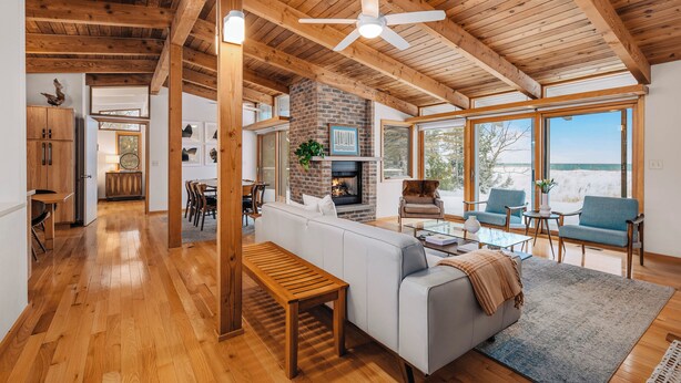 A spacious living area with a fireplace, wooden ceiling beams, and large windows offering a view of the outdoors. - Dune Ranch | Private Lake Michigan Beach + Fire Pit | Cathead Bay (Northport)