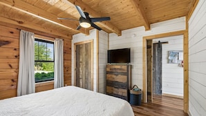Ein Schlafzimmer einer Holzhütte mit Bett, Deckenventilator, Fernseher und einem Fenster mit Vorhängen.