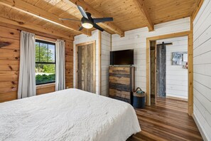 Camera da letto in una baita di legno, con un letto, un ventilatore a soffitto, una TV e una finestra con tende.