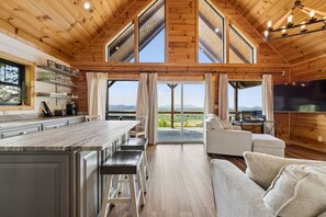 Un'ampia cucina con una grande isola, una zona pranzo con un tavolo alto e panche, un soggiorno con divano e TV, e ampie finestre che offrono vista sull'esterno.