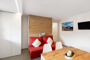 Een moderne kamer met een rode bank, witte stoelen, een houten tafel en een aan de muur bevestigde televisie.