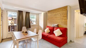 A modern living room with a wooden accent wall, a red sofa with white cushions, a dining table with chairs, and a large window with curtains. - Apartment 'A Due Passi Verde' with Mountain View, Balcony and Wi-Fi (Livigno)