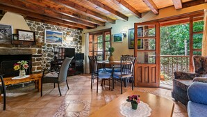 Una sala de estar acogedora con chimenea de piedra, vigas de madera en el techo, zona de comedor con sillas azules y ventanas amplias con vistas a la vegetación.