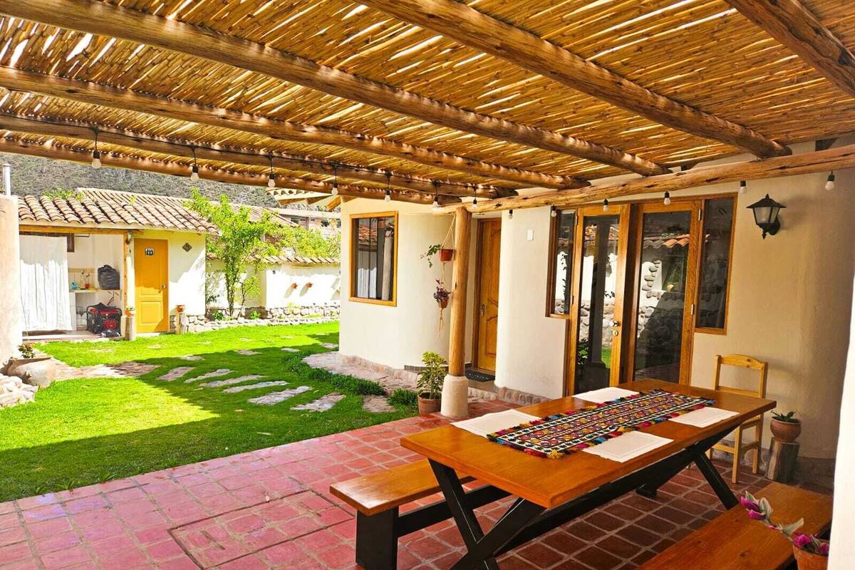 A covered patio area with a wooden table and benches, a small garden, and a building with a tiled roof in the background.