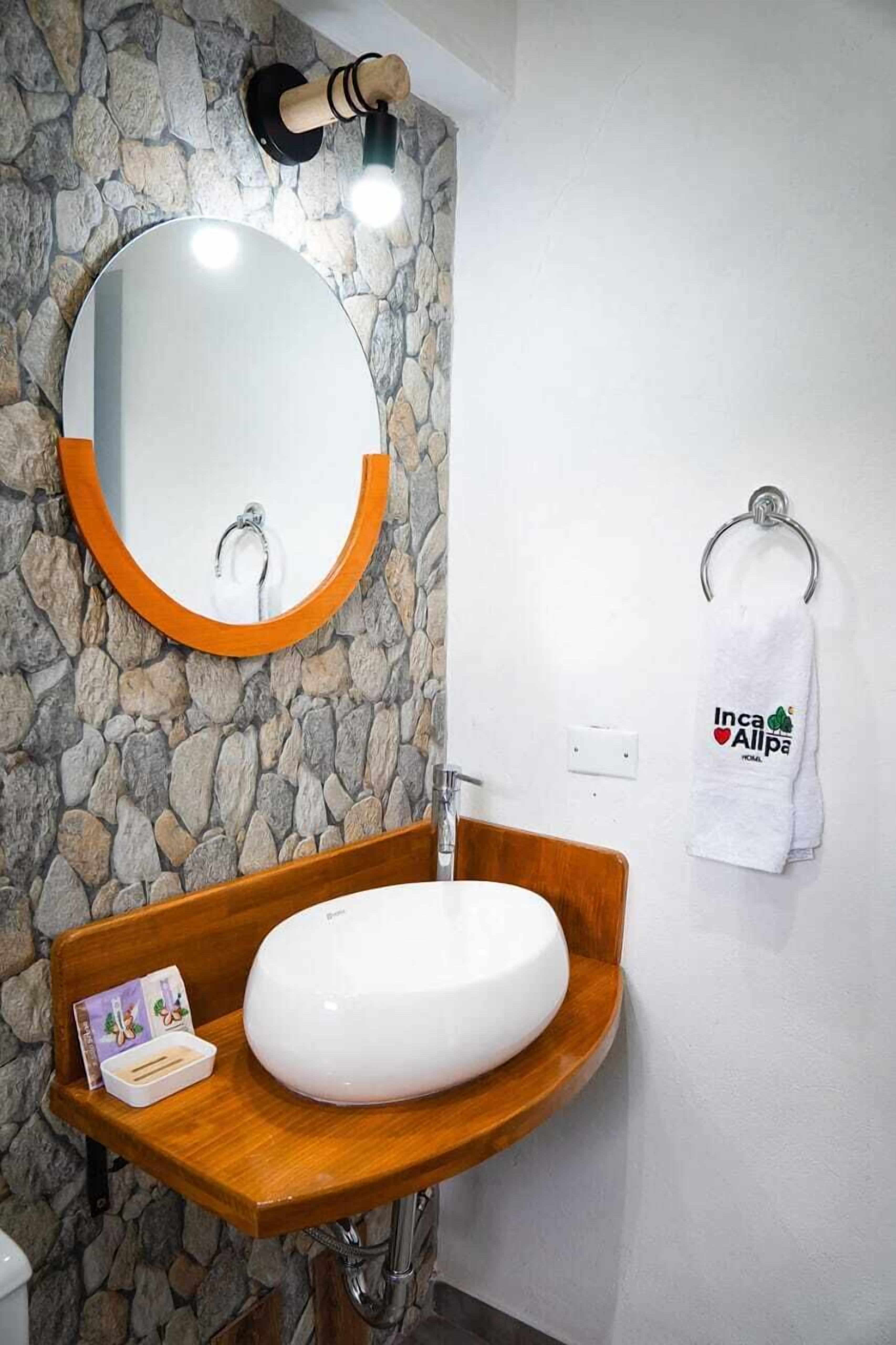 A bathroom with a stone wall, a round mirror with an orange frame, a wooden vanity, a white sink, and a towel rack with a towel.