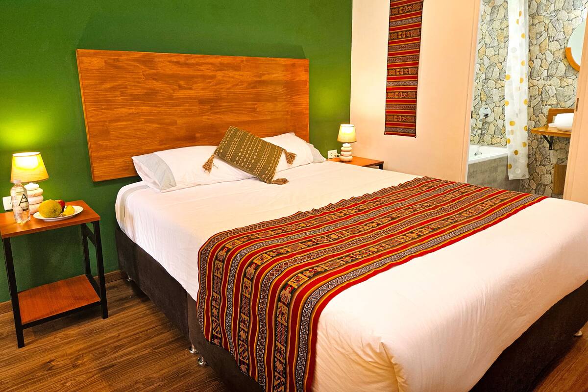 A neatly made bed with a patterned bedspread, a wooden headboard, and a bedside table with a lamp and a plate of fruit.