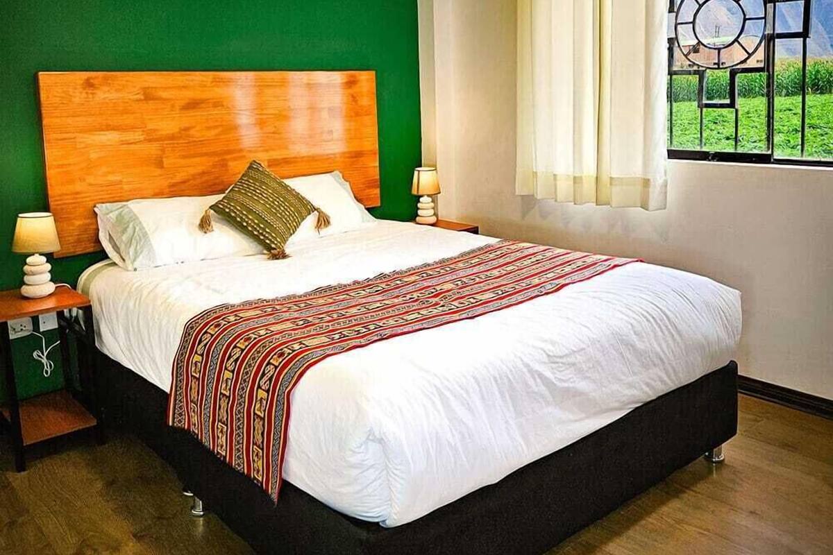 A bedroom with a wooden headboard, a large bed with white bedding, a red and black patterned throw, a nightstand with a lamp, and a window with a view of greenery and mountains.