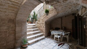 Un arc en pierre mène à une cour intérieure où se trouvent une table et des chaises, des plantes en pot et un escalier.
