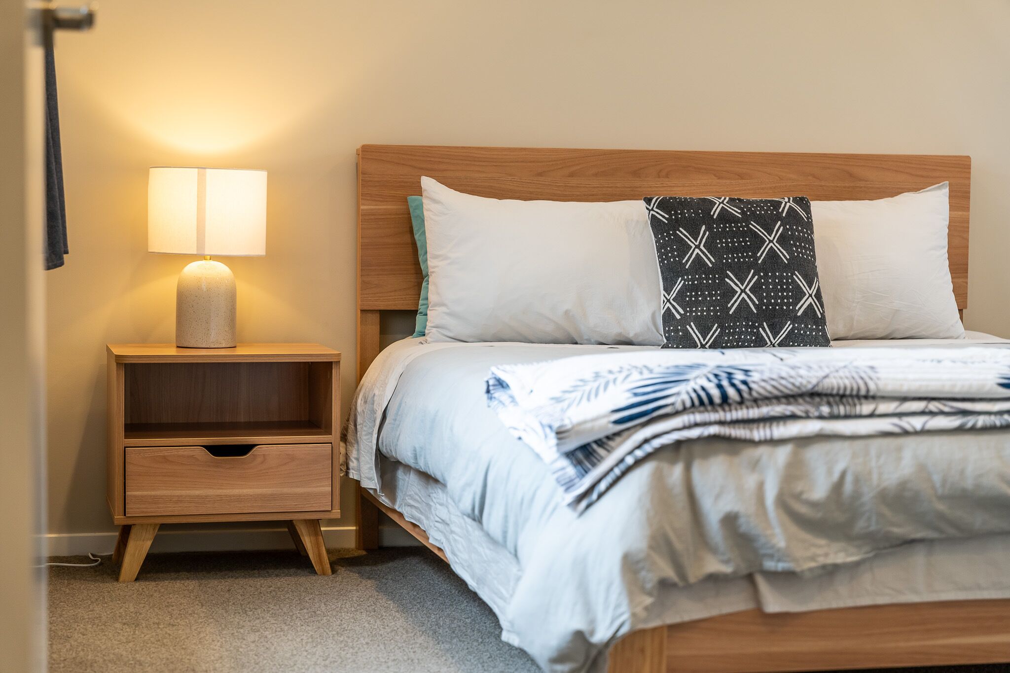 Een netjes opgemaakt bed met een bedsprei met patroon, een bedlampje en een nachtkastje van hout met een lade.