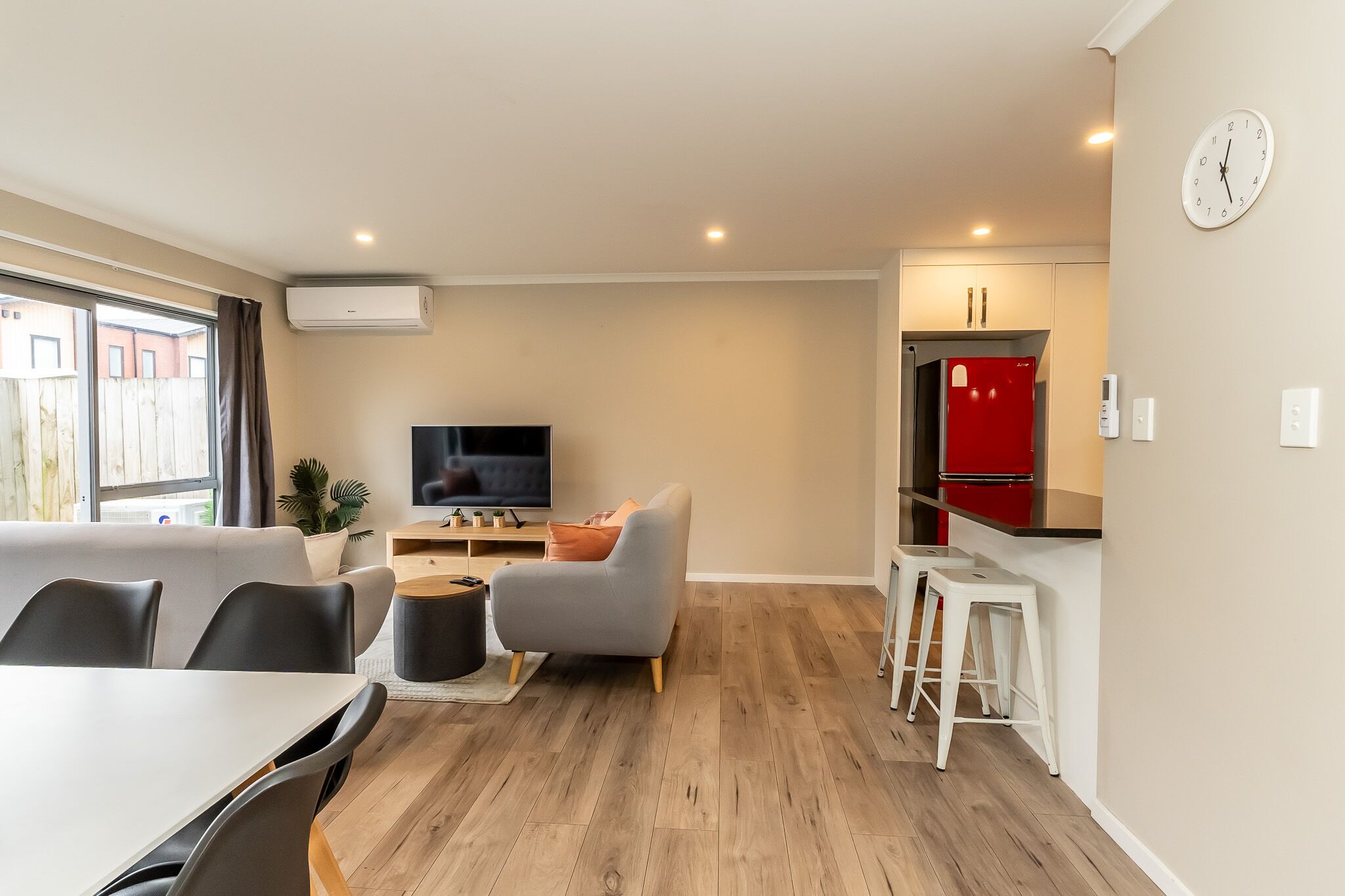 A modern living room with a flat-screen TV, grey sofa, dining table, and white bar stools.