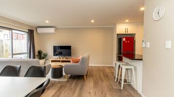 A modern living room with a flat-screen TV, grey sofa, dining table, and white bar stools.