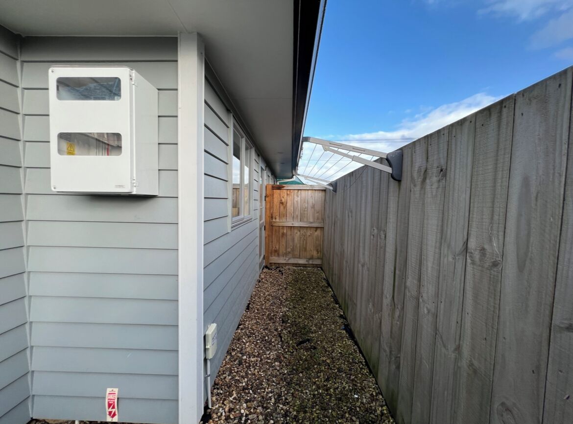 A narrow outdoor corridor with a wooden fence, a white utility box, and a gravel path.