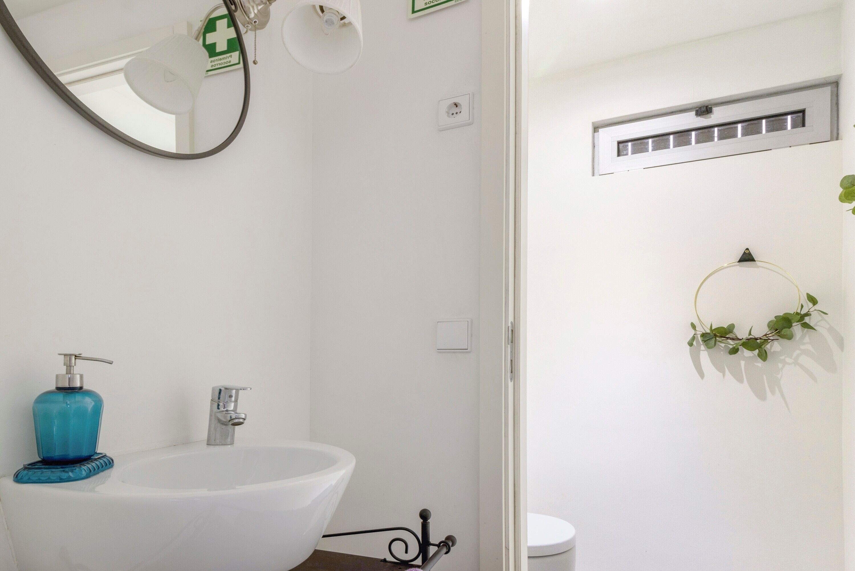 Ein Badezimmer mit einem weißen Waschbecken, einem blauen Seifenspender, einem Spiegel, einer Lampe, einer Zimmerpflanze und einer weißen Toilette.