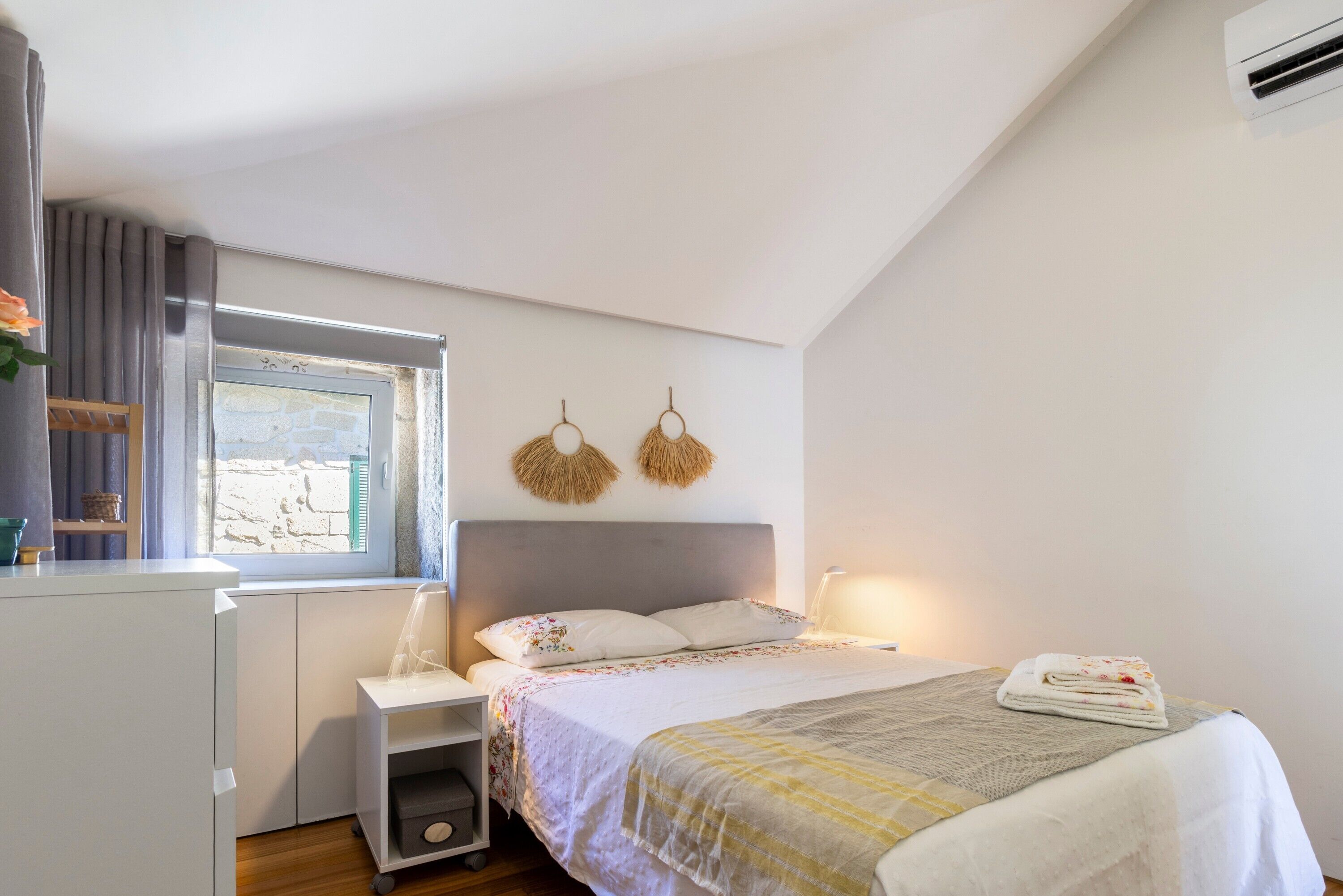 Una camera da letto con un letto, un comodino, una finestra con tende, un condizionatore d'aria a muro e una piccola mensola con un cesto.