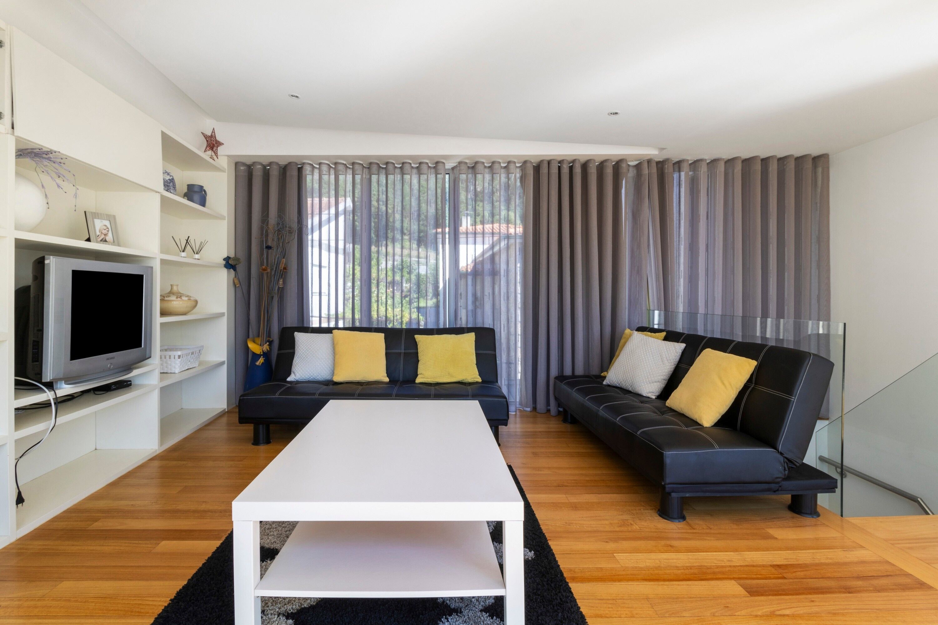 Ein modernes Wohnzimmer mit einem weißen Couchtisch, zwei schwarzen Sofas mit gelben Kissen, einem Flachbildfernseher und einem großen Fenster mit grauen Vorhängen.