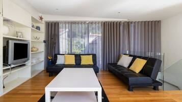 Ein modernes Wohnzimmer mit einem weißen Couchtisch, zwei schwarzen Sofas mit gelben Kissen, einem Flachbildfernseher und einem großen Fenster mit grauen Vorhängen.