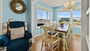 A dining area with a wooden table and white chairs, a blue wall, and a view of the ocean. - Escape by the Sea! NEW! (Galveston)