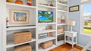 A well-organized room with white shelving, wicker baskets, a television, a small table with a lamp, and a window with a view. - Escape by the Sea! NEW! (Galveston)