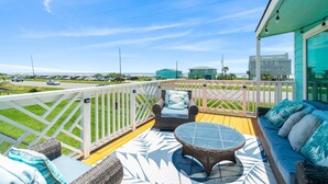 A deck with wicker furniture, a glass table, and a view of a road and buildings. - Escape by the Sea! NEW! (Galveston)