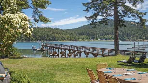 Terraza junto al lago con una mesa y sillas, con vista a un lago tranquilo con barcos y un muelle de madera.