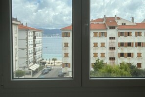 Udsigt gennem et vindue af en kystbygning med flere etager og altaner, samt en gade foran.