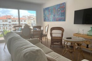 En stue med sofa, stole, et bord, et fjernsyn og en balkon med udsigt til bygninger.