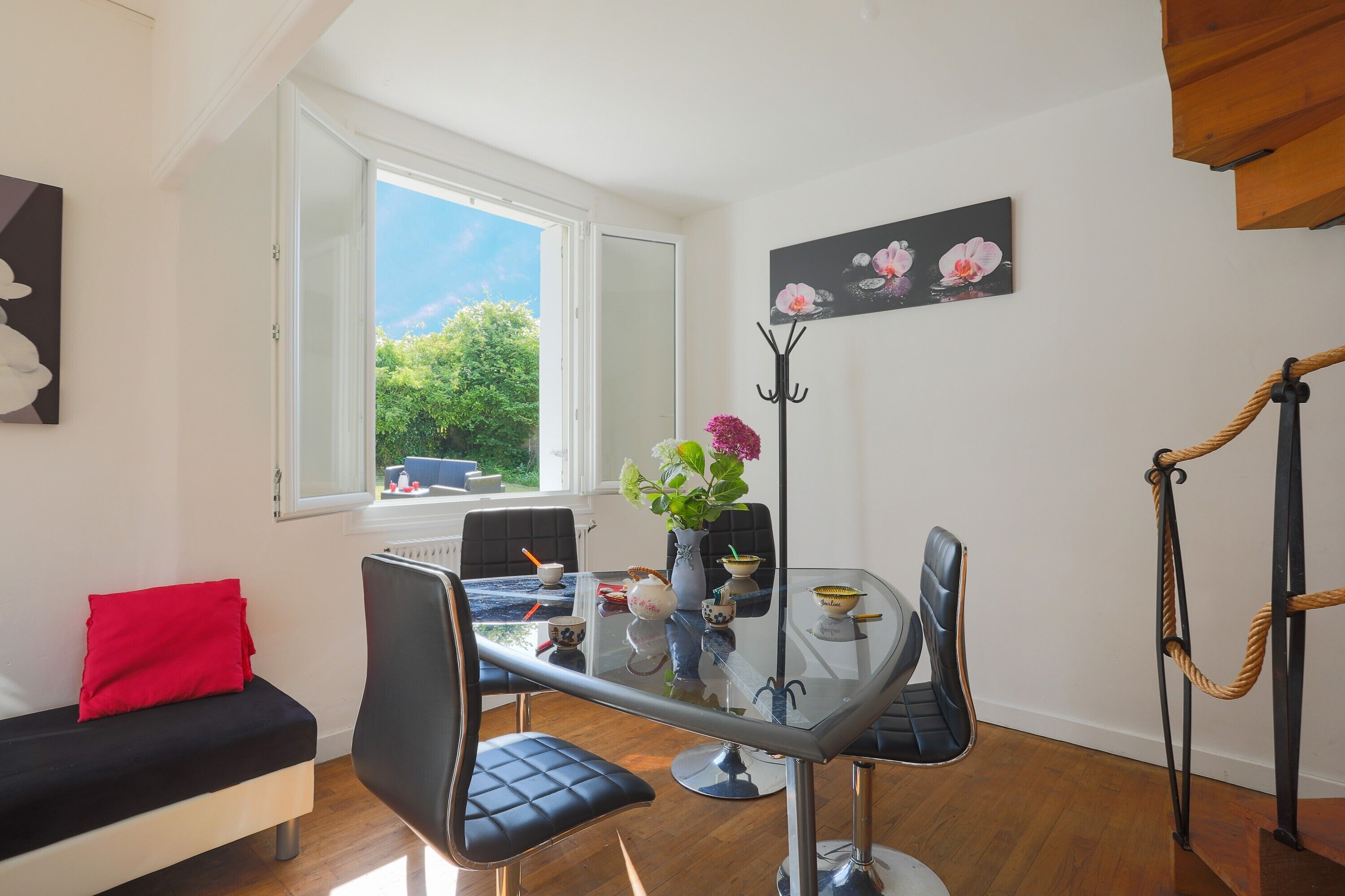 A modern room with a glass dining table, black leather chairs, a red cushion on a black sofa, a vase with flowers, and a painting of flowers on the wall.