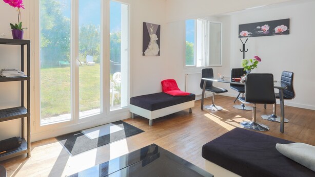 A modern living room with a sofa, a dining table with chairs, a small table with a chair, and a large window with a view of the outdoors. - Holiday Home 'Maison Bretonne' with Private Terrace, Private Garden and Balcony (Ploumagoar)