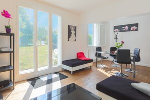 A modern living room with a sofa, a dining table with chairs, a small table with a chair, and a large window with a view of the outdoors. - Holiday Home 'Maison Bretonne' with Private Terrace, Private Garden and Balcony (Ploumagoar)