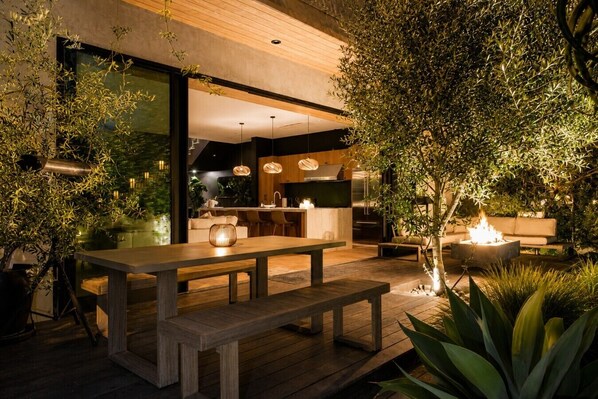 An outdoor dining area with a wooden table and benches, surrounded by greenery and a fire feature.
