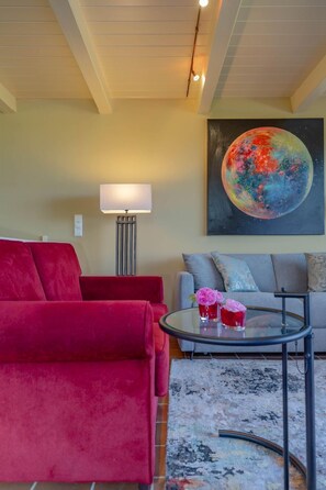 Uma sala de estar com um sofá vermelho, um sofá cinzento, uma mesa de centro de vidro com flores cor-de-rosa, uma luminária e um quadro de um planeta na parede.
