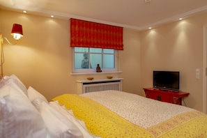 Um quarto com uma cama, uma janela com bajela vermelha, um radiador abaixo da janela, uma televisão sobre suporte vermelho e um abajur fixado na parede.