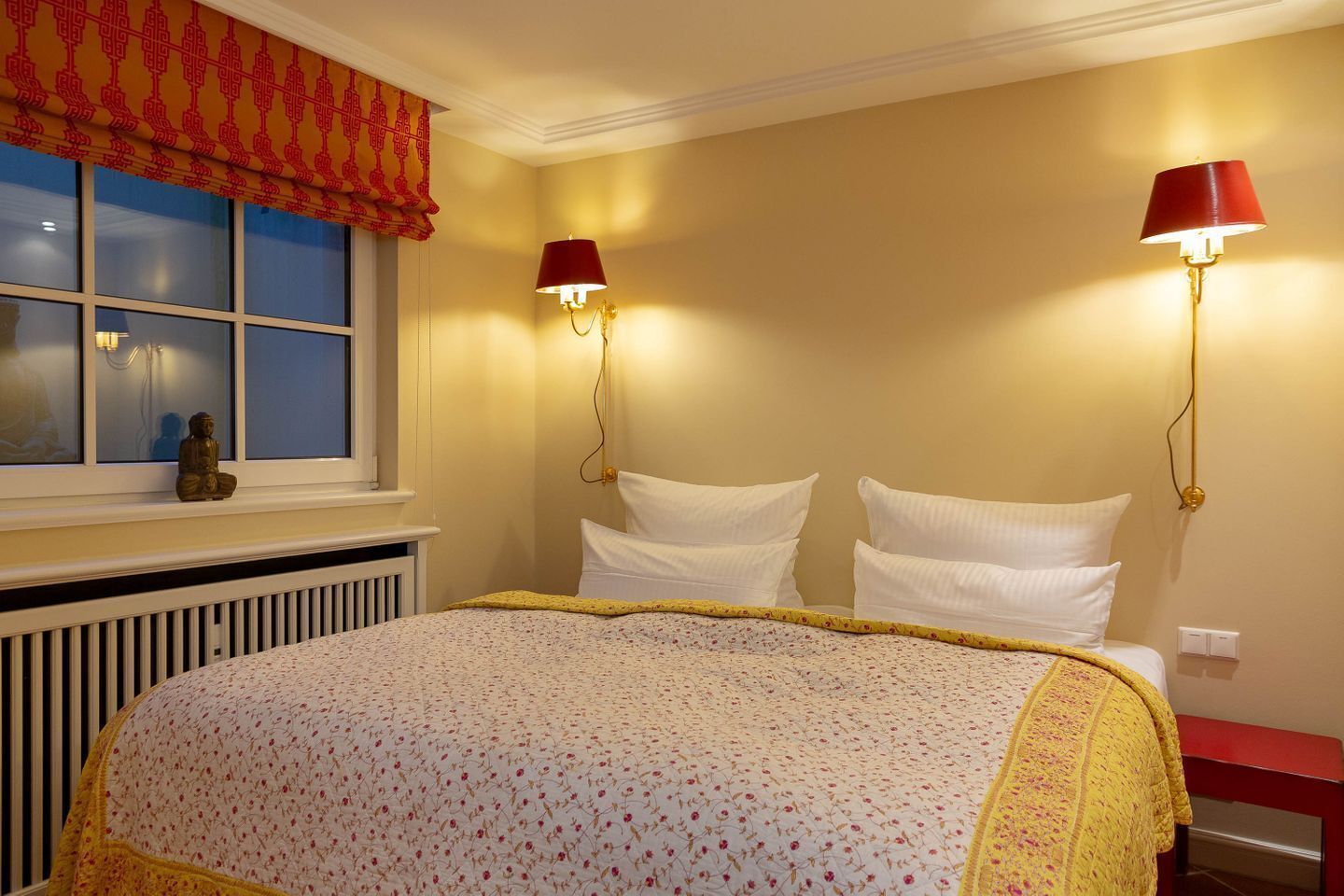 Een slaapkamer met een bed, twee bedlampjes, een raam met een rode vouwgordijn en een radiator onder het raam.