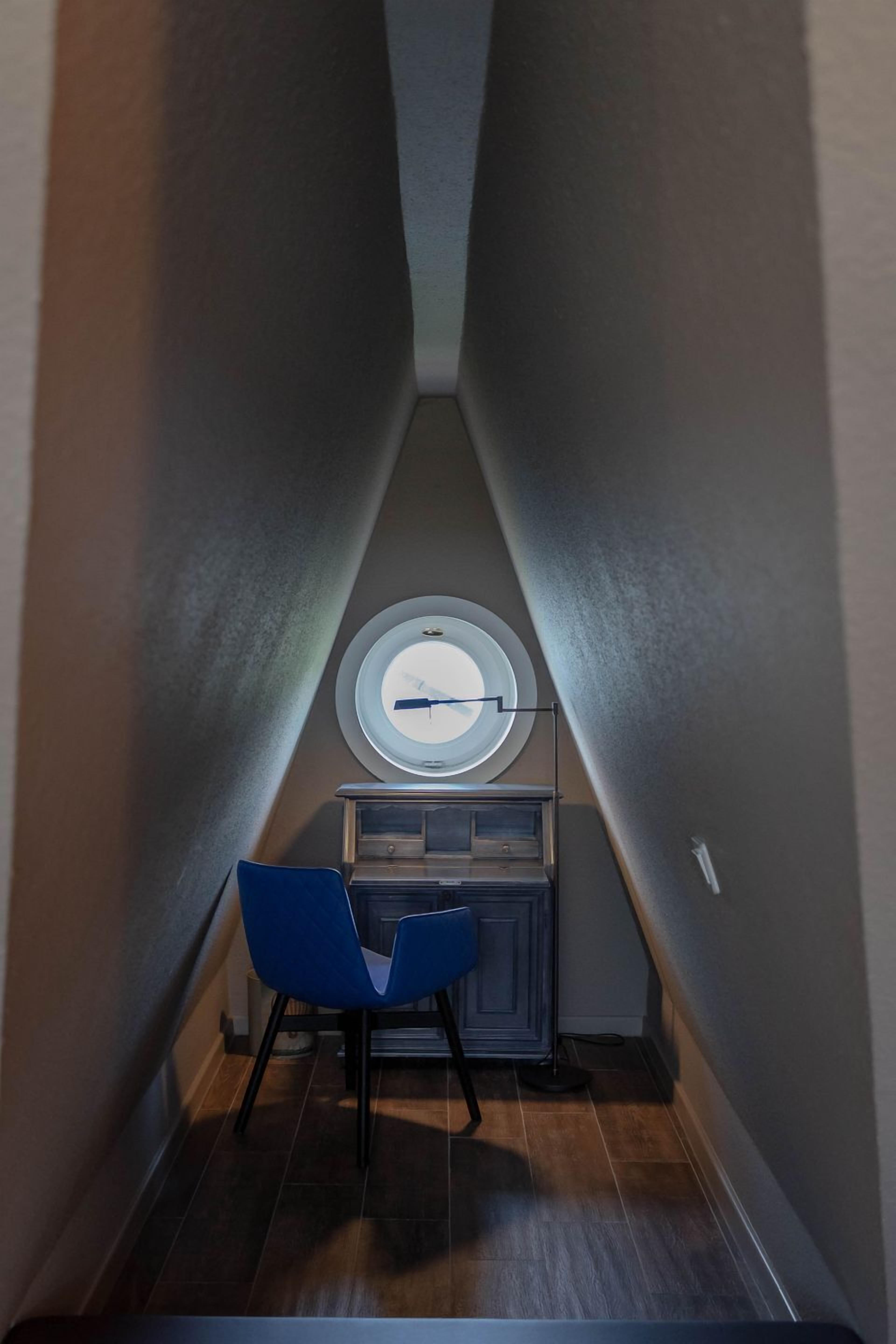 A narrow hallway with a blue chair, a small table, and a round window with a light fixture.