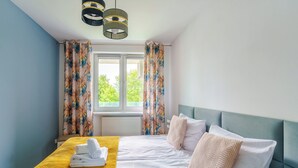 Una cama bien tendida con una colcha amarilla y almohadas blancas, una ventana con cortinas y una pared azul.