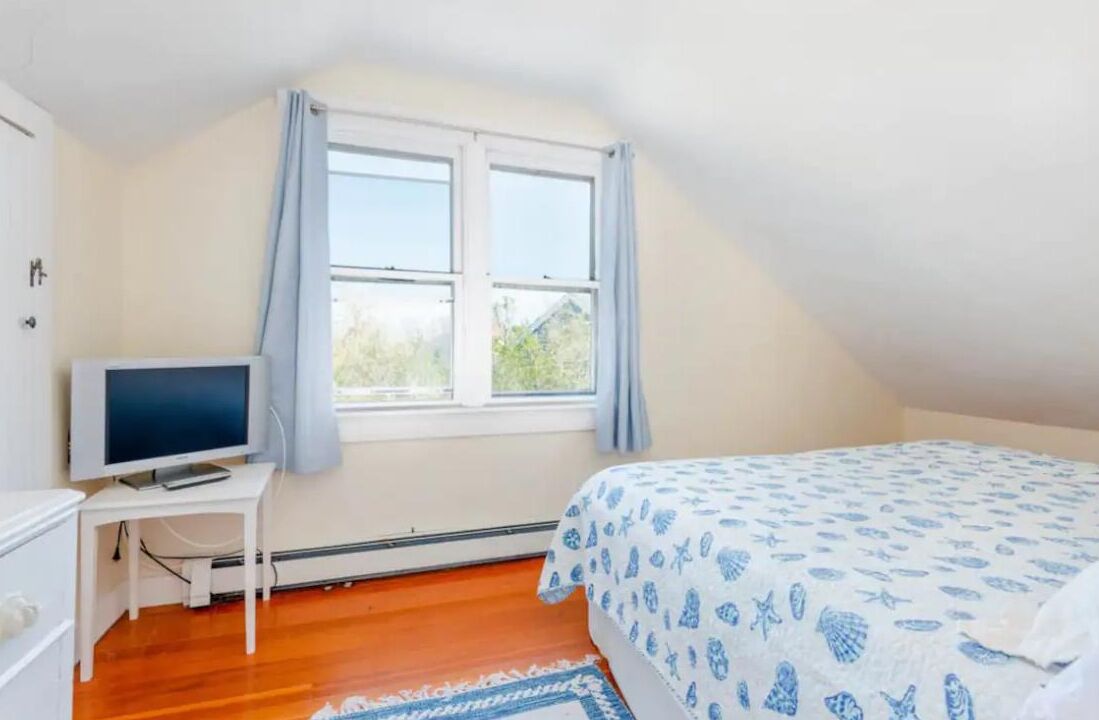 Quarto com cama, televisão sobre uma mesinha, janela com cortinas e vista para o verde lá fora.
