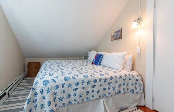 A small, cozy bedroom with a sloped ceiling, a bed with a nautical-themed bedspread, a single wall-mounted night light, and a small wooden stool. - Cozy 4 bedroom cottage in town, with parking  (Nantucket)