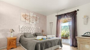 A bedroom with a bed, bedside tables, a lamp, a decorative headboard, a window with curtains, and a view of greenery outside.