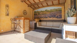 A room with a stone wall, a wooden ceiling, a mural of a vineyard, a white sofa, a bar area with stools, and a cactus plant.