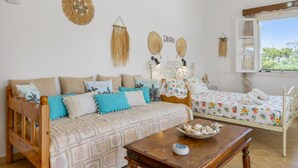 Une pièce comprenant un canapé en bois, un lit plus petit, une table agrémentée d’objets décoratifs, ainsi que des éléments de décoration murale, notamment une œuvre en macramé et deux ventilateurs fixés au mur.